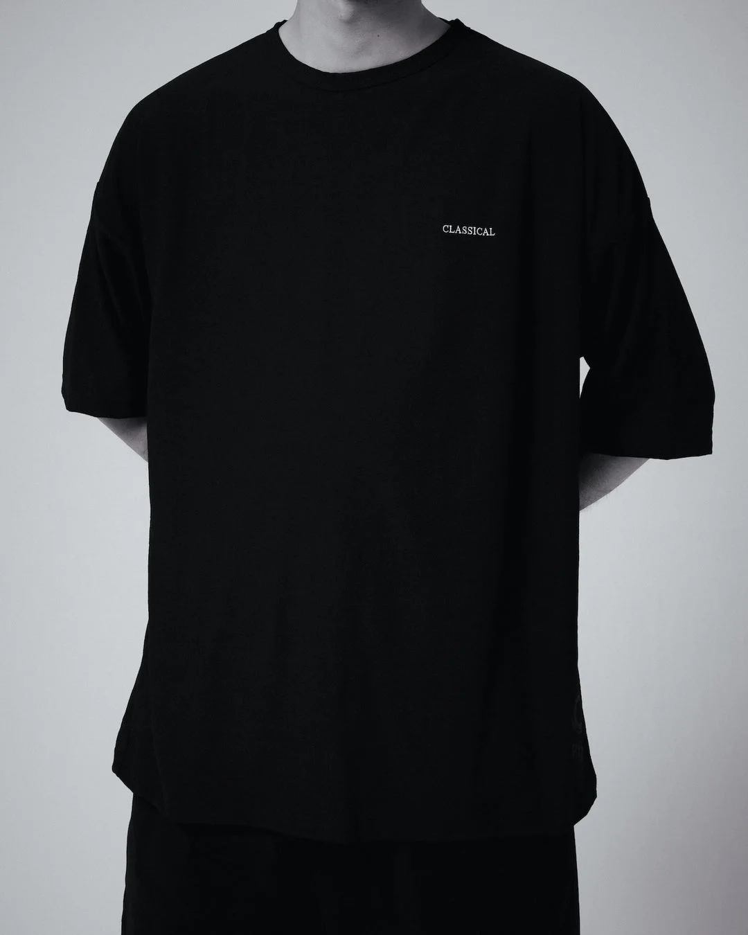 「CLASSICAL」の白いロゴが入った黒いオーバーサイズTシャツを着用した人物の、胸元から腰部を写した画像です。シンプルでモダンなデザインが特徴のファッションアイテムです。