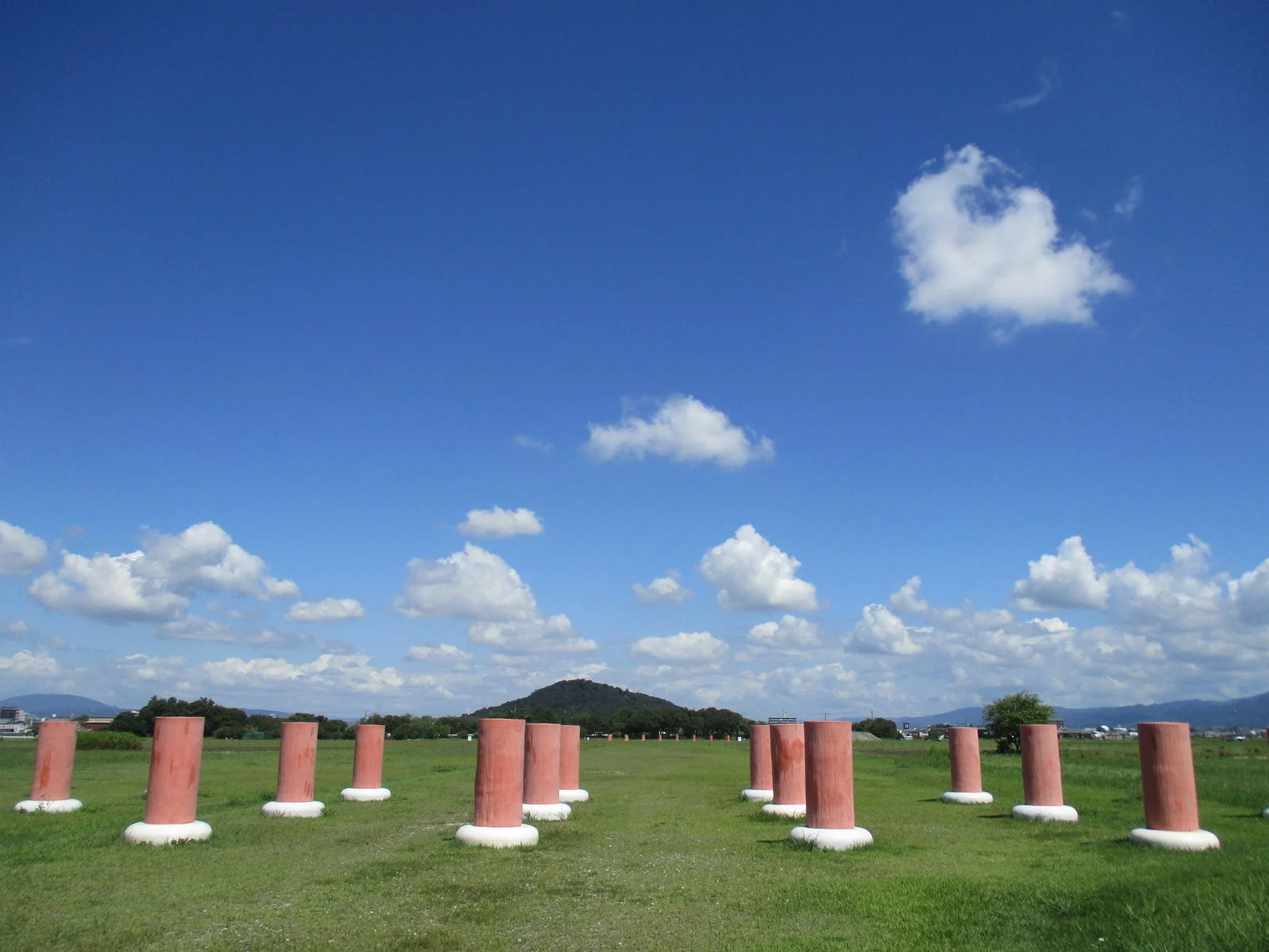 青い空と白い雲が広がる開放的な草原に、赤茶色の円筒形の柱が点々と並んでいる風景