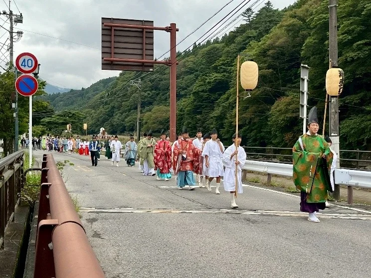 山間部の道を、伝統的な衣装をまとった人々が提灯を持って歩く行列の様子