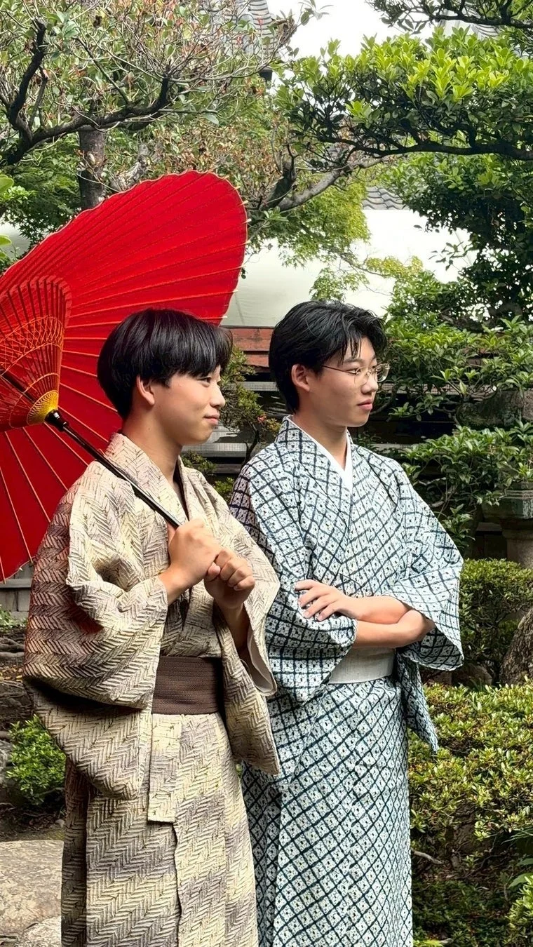 日本庭園で和服（浴衣）を着た若い男性2人が並び、1人は赤い和傘を携えています