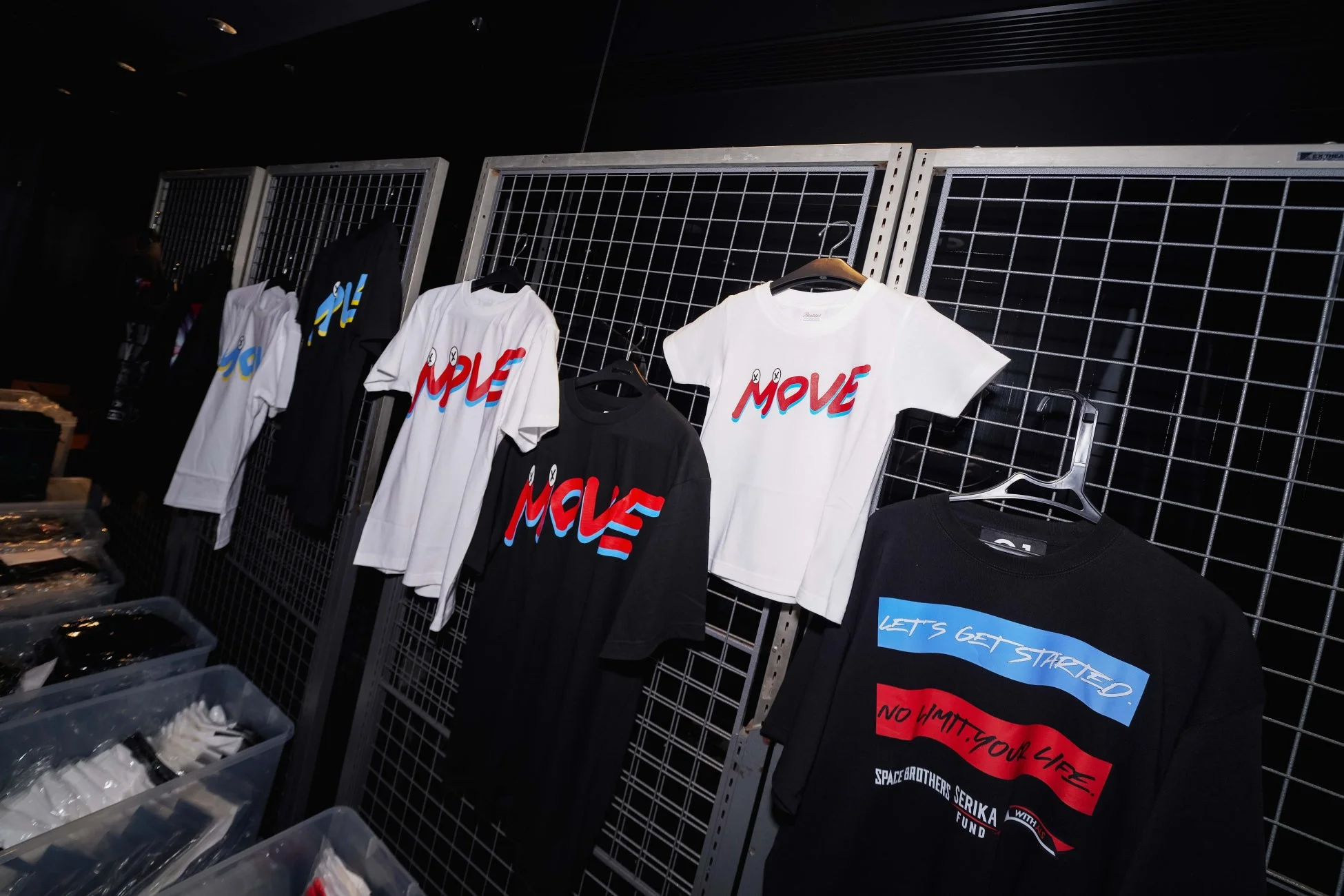 暗い空間に金属製のディスプレイラックが並び、複数のTシャツが展示されています。白と黒のTシャツには「MOVE」の文字が、別の黒いTシャツには「LET'S GET STARTED.」や「NO LIMIT, YOUR LIFE.」などのメッセージ、さらに「SPACE BROTHERS SERIKA FUND」の文字が見えます。これはアパレル商品の販売または展示会の様子を捉えたものです。