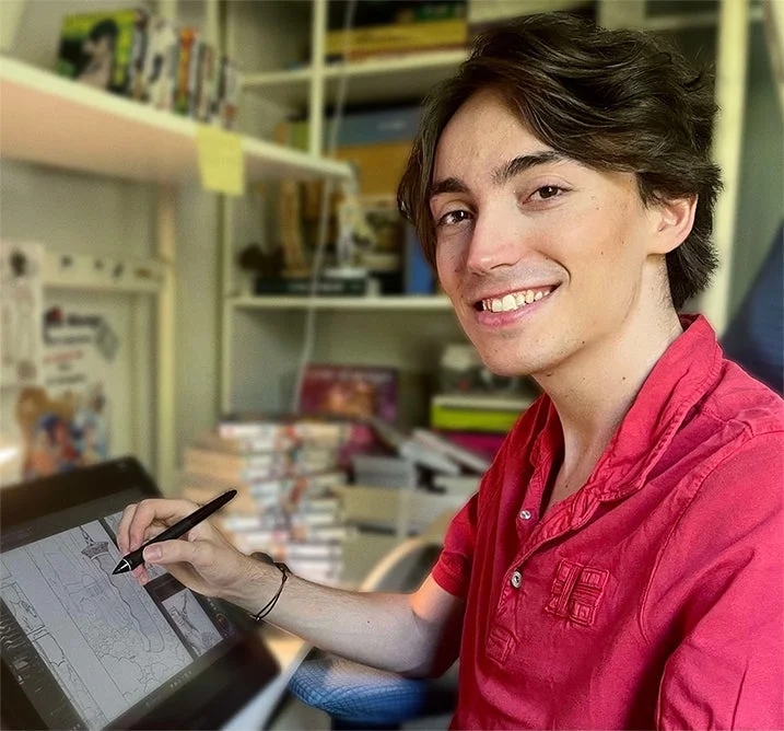 赤いポロシャツを着た若い男性が、デジタルペンを使ってペンタブレットで絵を描いている。笑顔でカメラを見つめており、クリエイティブな作業に集中している様子がうかがえる。背景には棚に並んだ本が見える。
