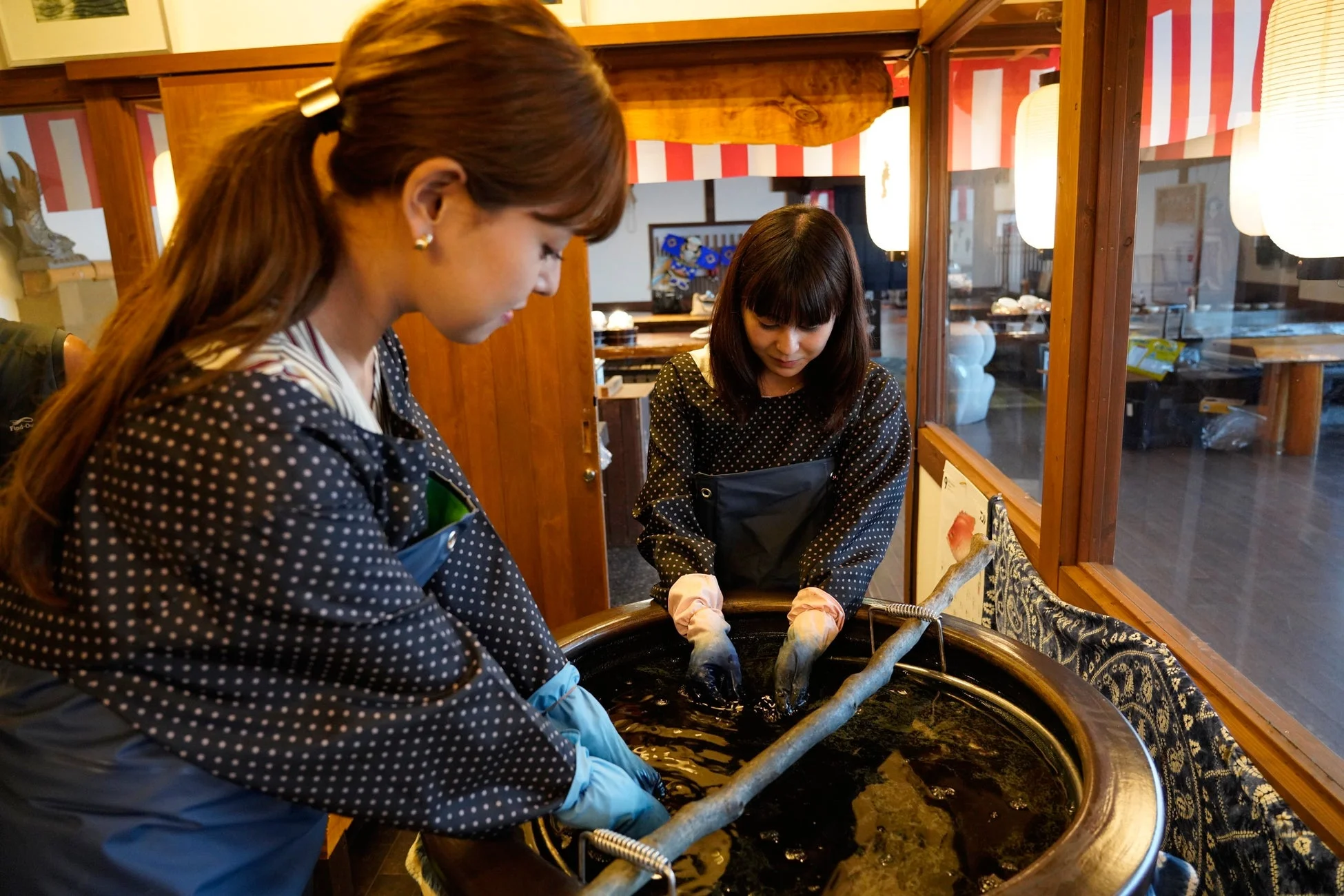 二人の女性が伝統的な染物体験をしている様子。エプロンと手袋を着用し、大きな桶の中で布を染めている。和風の店内で集中して作業に取り組んでいる。