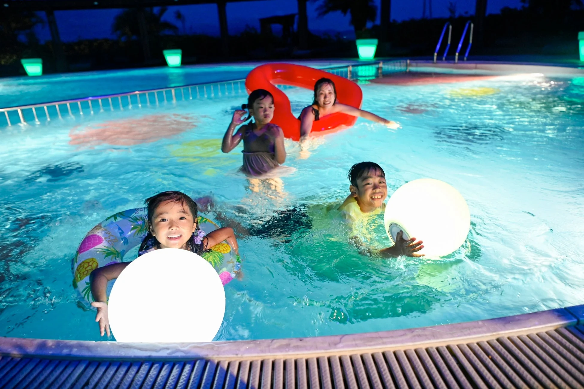 夜のプールで楽しむ家族の様子が写っています。大人1人と子供3人が水着で泳ぎ、浮き輪や光るボールで遊んでいます。プールは青くライトアップされ、皆笑顔で楽しいひとときを過ごしているようです。