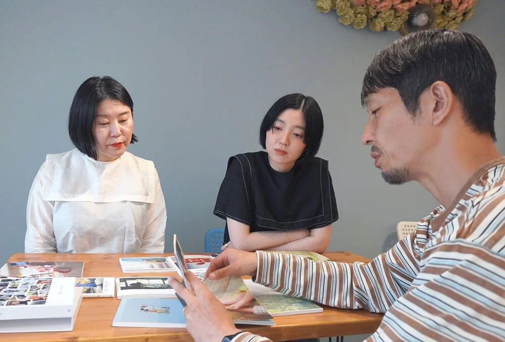 木製のテーブルを囲んで座る3人(男性1人、女性2人)が、本や写真集を見ながら話し合っている様子