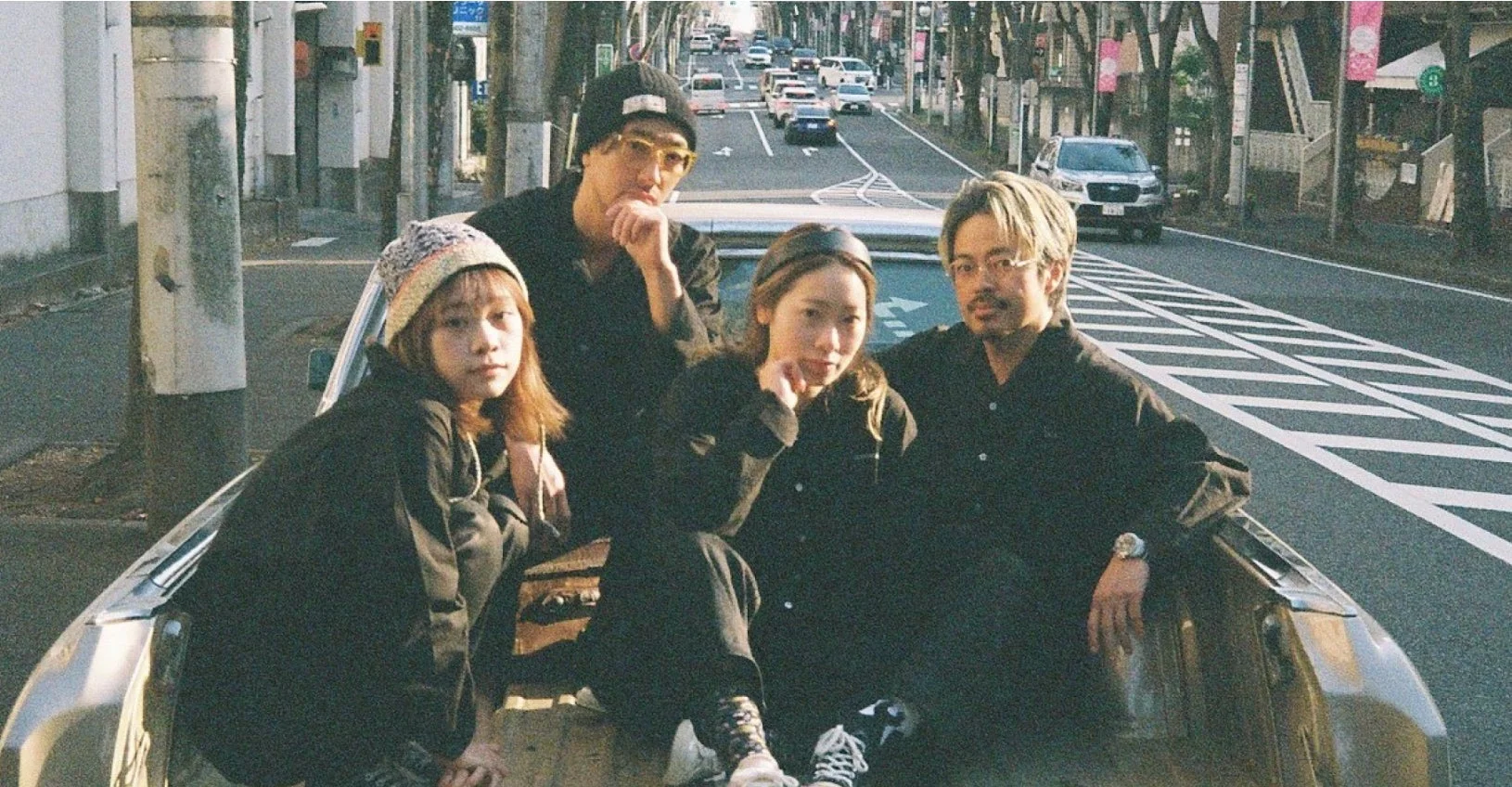 4人の男女が車の荷台に座り、カメラに向かってポーズをとっているストリートスナップのような写真です。都市の道路を背景に、全員がダークトーンの服を着用し、クールでリラックスした雰囲気が漂っています。