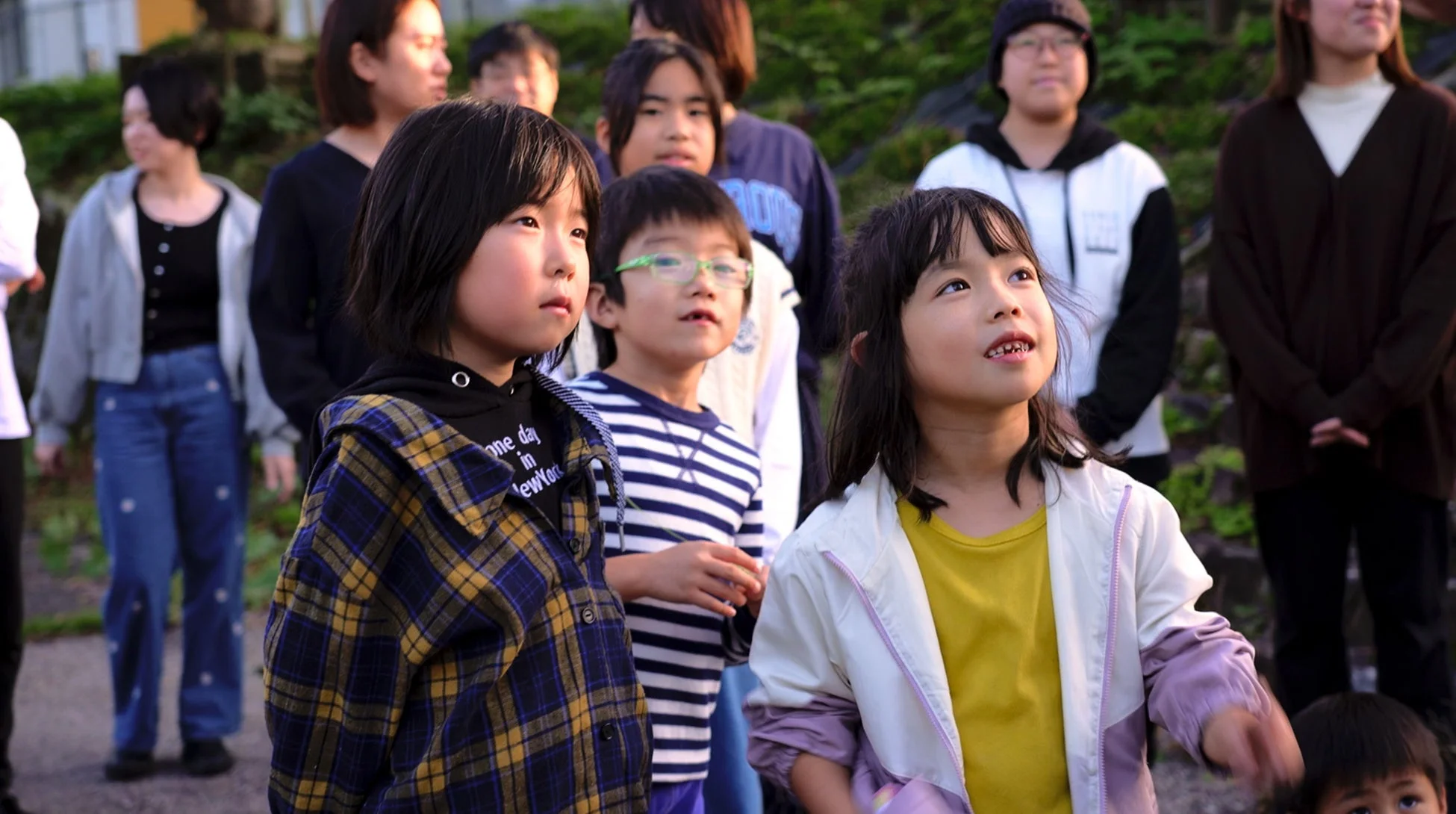 屋外で集まった子供たちが、空や遠くの何かを見上げている様子を捉えた写真です。好奇心に満ちた表情が印象的で、周囲には大人たちも見守っています。和やかな雰囲気のイベントの一場面のようです。