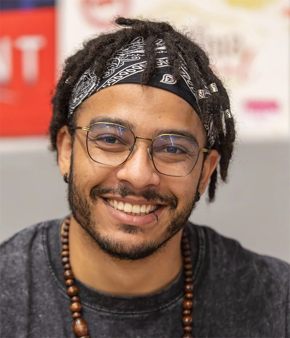ドレッドヘアにバンダナと眼鏡をかけた男性が、明るい笑顔を見せているポートレート。唇にはピアスがあり、木製のビーズネックレスを着用している。