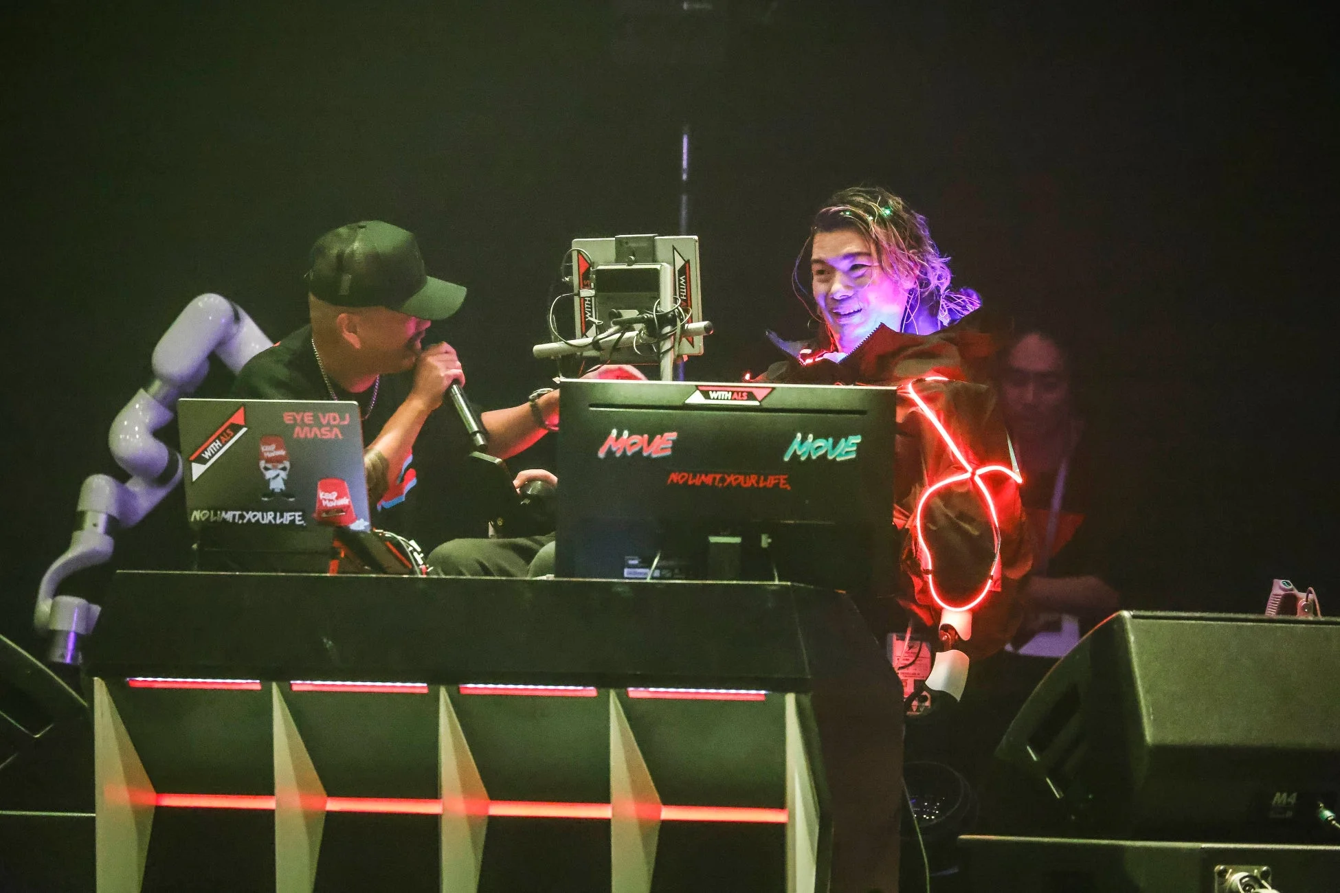ステージ上でDJ/VDJがパフォーマンスしており、マイクを持つ人物とネオンライトを身につけた人物が映っています。機材には「MOVE」や「NO LIMIT, YOUR LIFE.」、「WITH ALS」などの文字が見られ、背景にはロボットアームも確認できます。ライブ感のあるエネルギッシュなシーンです。