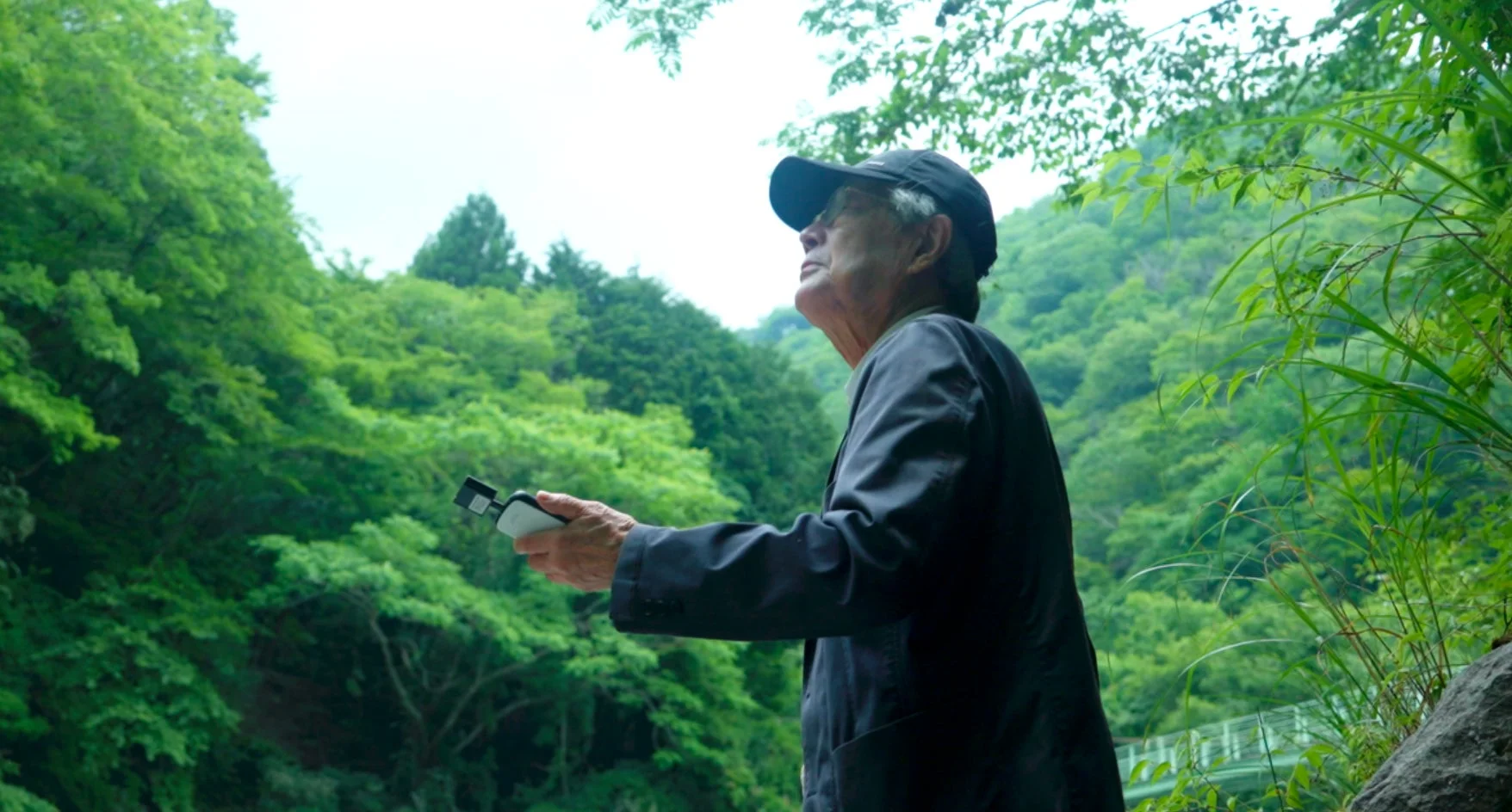 豊かな緑に囲まれた自然の中で、帽子をかぶった高齢の男性がデバイスを手に持ち、上空を見上げている様子。落ち着いた雰囲気で自然を満喫しているようだ。