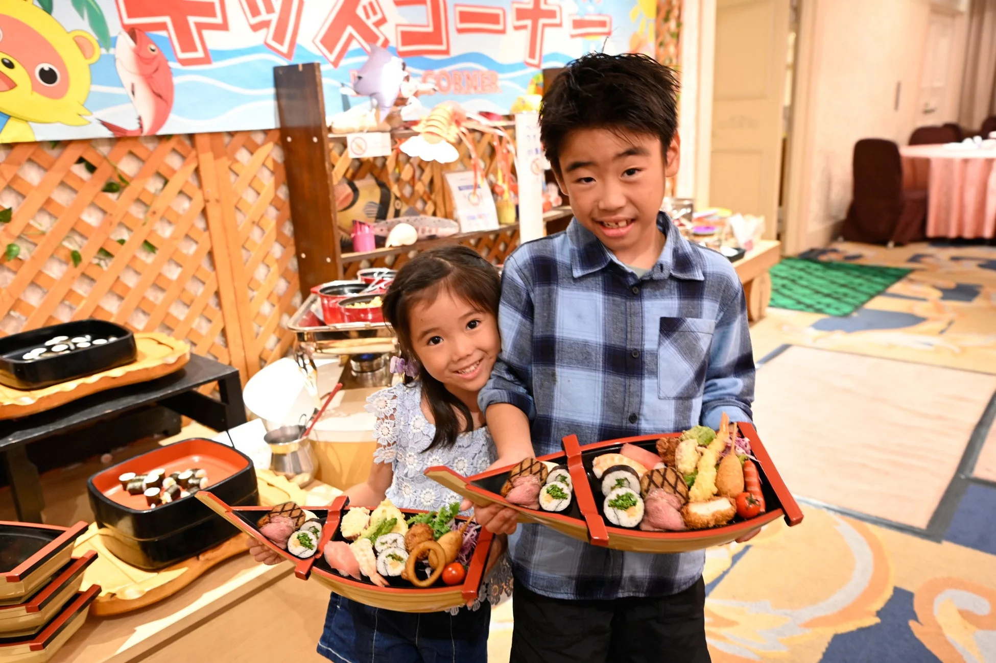 キッズコーナーで、男の子と女の子が笑顔で船盛りの寿司や天ぷらなどの和食を持っている様子です。奥にはビュッフェ形式の料理が並んでいます。