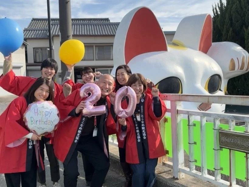 とこなめ観光協会のハッピを着た人々が、大きな招き猫の像の前で笑顔で集まっています