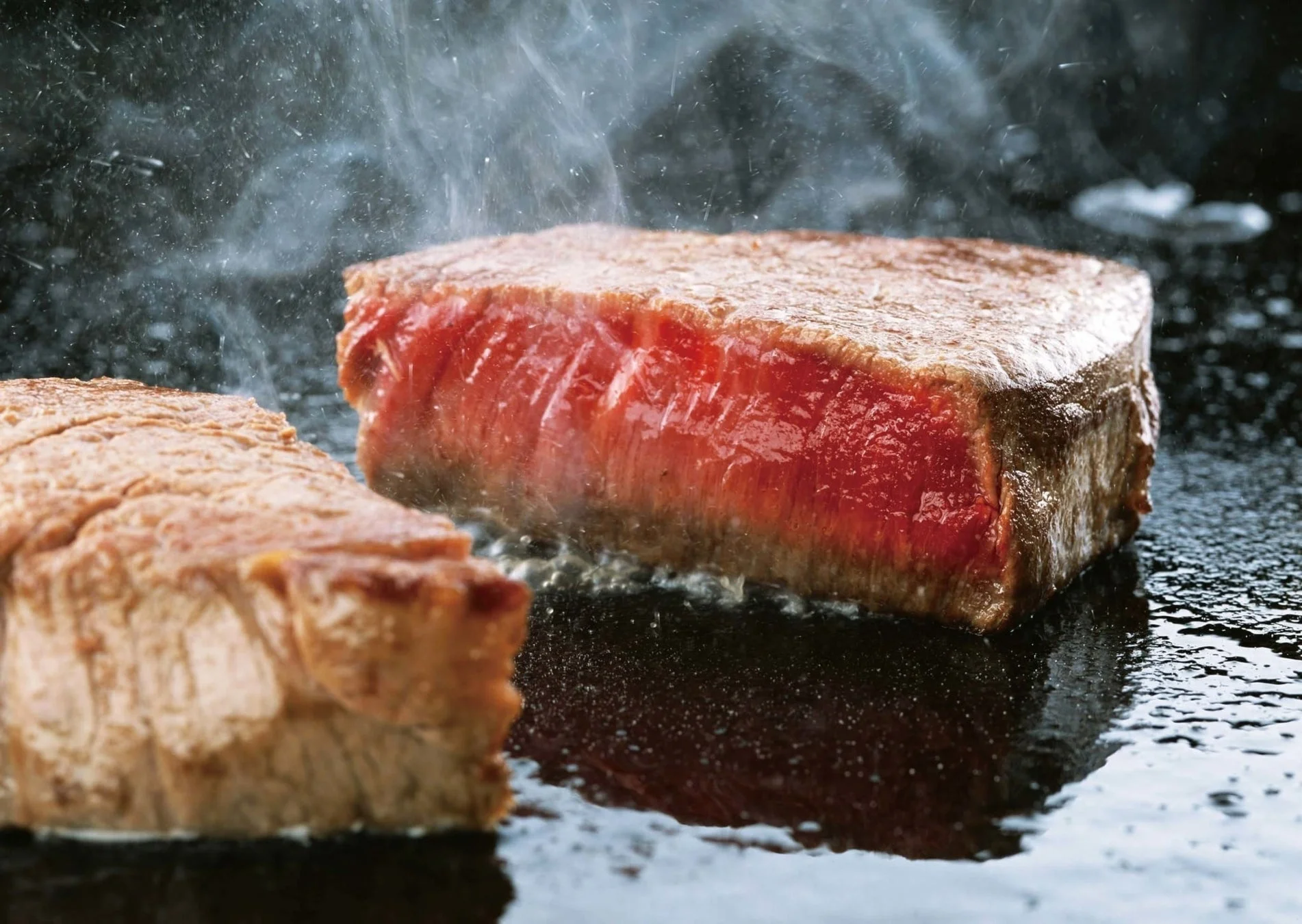 熱い鉄板の上で分厚いステーキ肉が焼かれている様子を捉えた画像です。表面は香ばしく焼け、中は鮮やかな赤色を保っており、湯気と油の飛び散る様子が食欲をそそります。