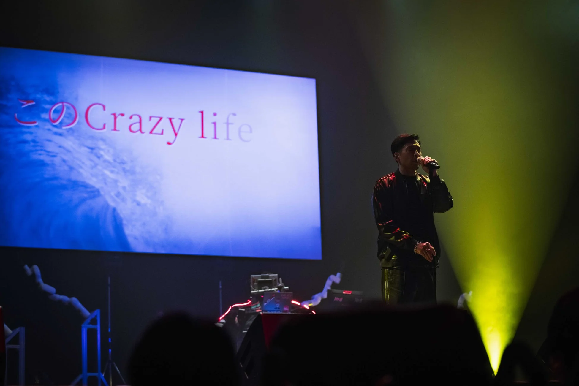 ステージ上で男性がマイクを持って歌っており、背後の大型スクリーンには「この Crazy life」という文字と青い波のような画像が映し出されています。黄色いスポットライトが印象的なライブパフォーマンスの様子です。