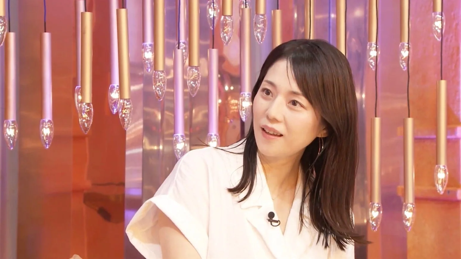 白いシャツを着た女性が、背景に多数の吊り下げ式照明があるスタジオのような場所で、マイクを付けて穏やかな表情で話している様子