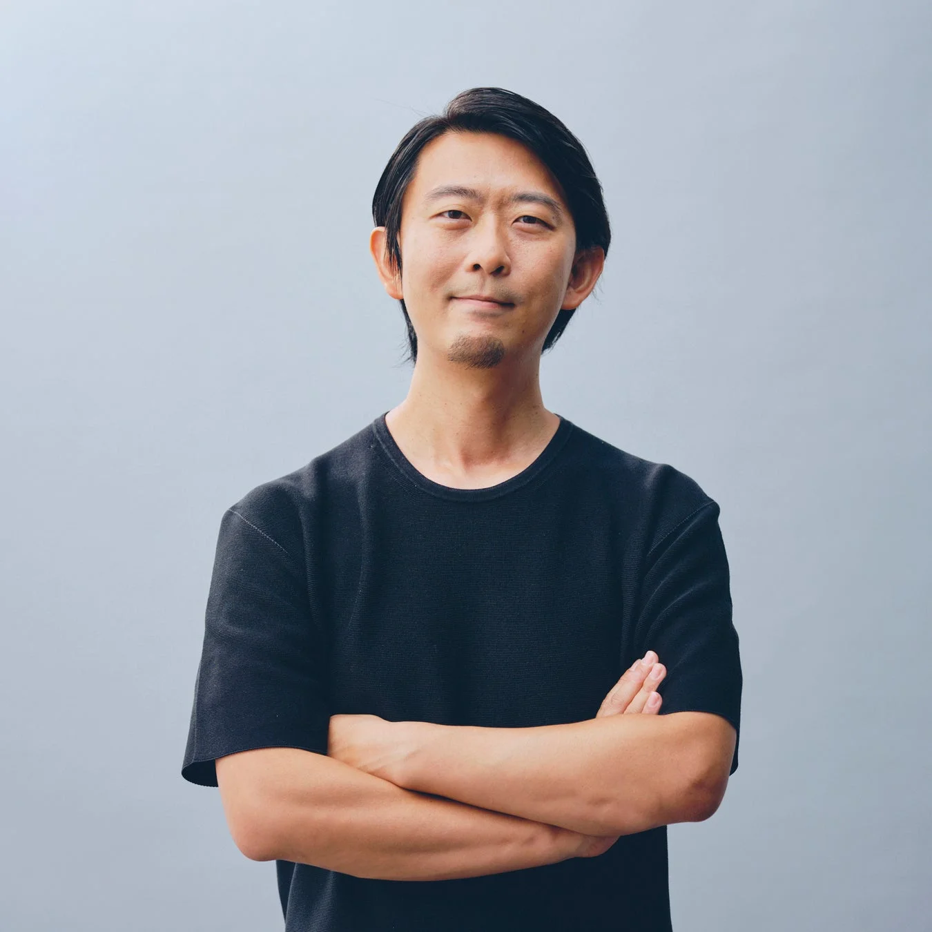 薄い灰色の背景を背に、黒いTシャツを着たアジア人男性が腕を組み、穏やかな笑顔でカメラを見つめている上半身のポートレート
