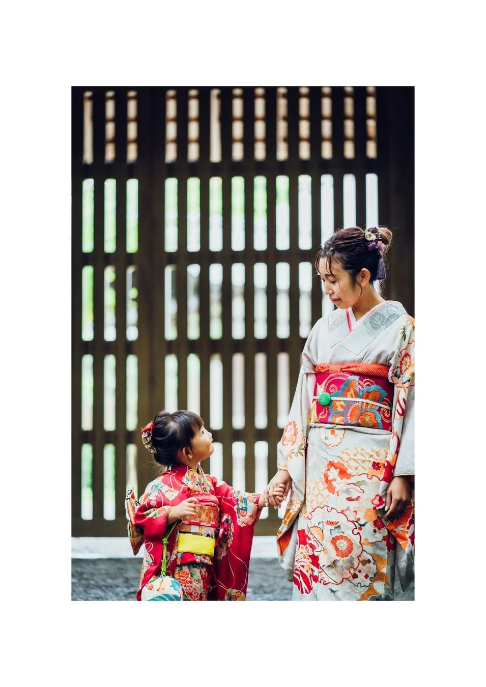 日本の伝統的な着物を着た女性と幼い女の子が手をつなぎ、木製の格子の前で立つ