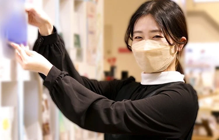 ベージュのマスクと黒い服を着た若い女性が、棚の商品に手を伸ばしながら笑顔を見せている様子