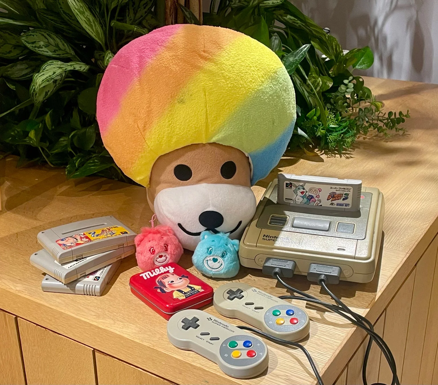 スーパーファミコン本体とコントローラー、複数のゲームソフト、カラフルな大きなぬいぐるみ、ケアベアの小さなぬいぐるみ、ミルキーの缶が並べられた、レトロゲームとキャラクターグッズのコレクションの様子