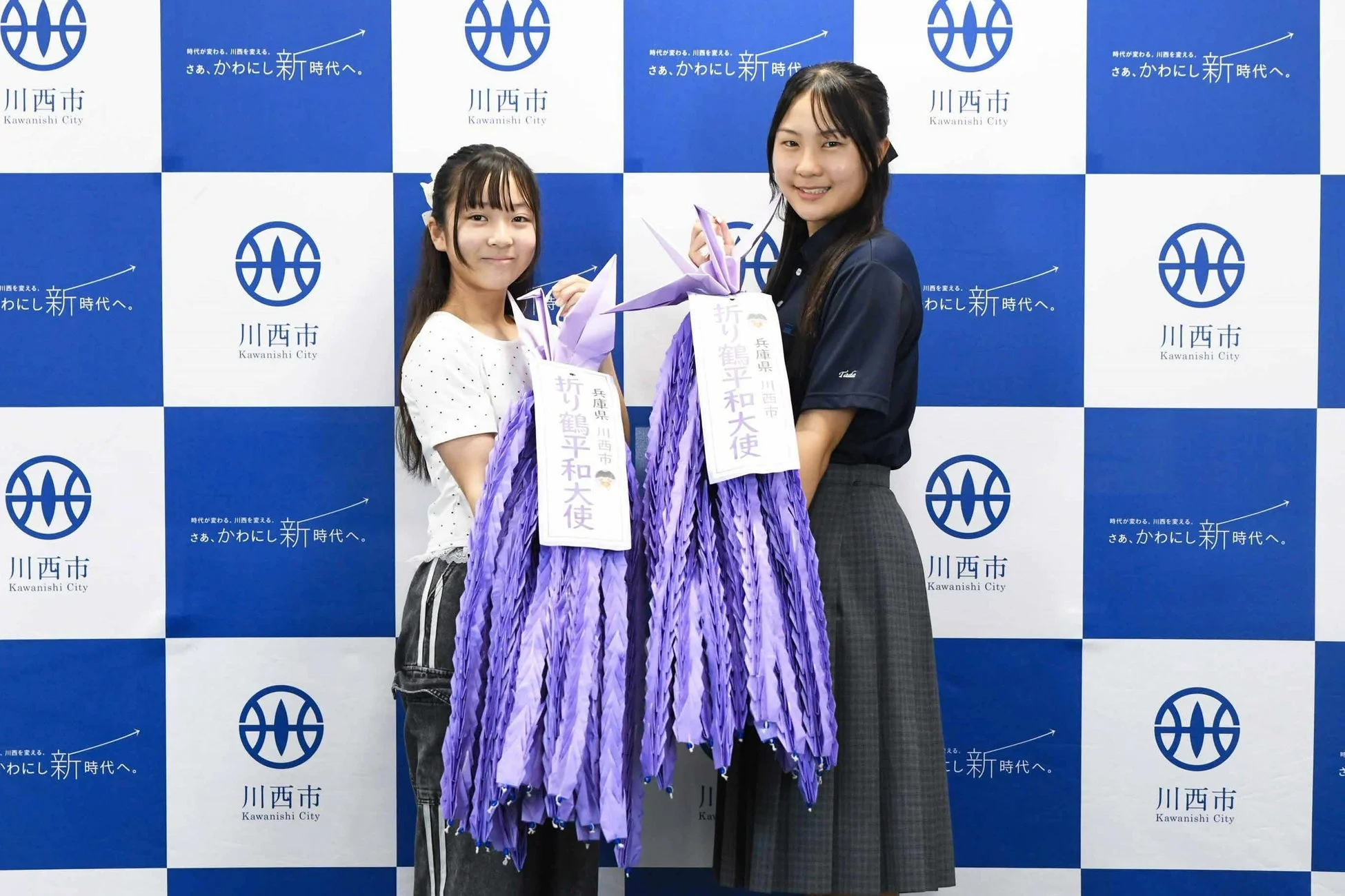 「折り鶴平和大使」に任命された川西市の若い女性二人が、市章とスローガンが描かれた背景の前で、紫色の折り鶴の束を手に笑顔を見せている。