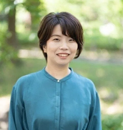 屋外で笑顔を見せるショートヘアのアジア人女性のポートレートです。彼女は青緑色のシャツを着用しており、背景は緑豊かな自然がぼかされています。
