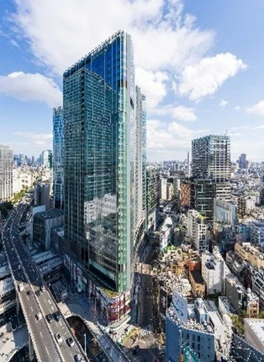 晴れた日に撮影された、高層ビルが立ち並ぶ近代的な都市の景観です。手前にはガラス張りの巨大なビルがあり、その横には車が行き交う高速道路が見えます。広がる街並みと青空に浮かぶ白い雲が印象的な一枚です。