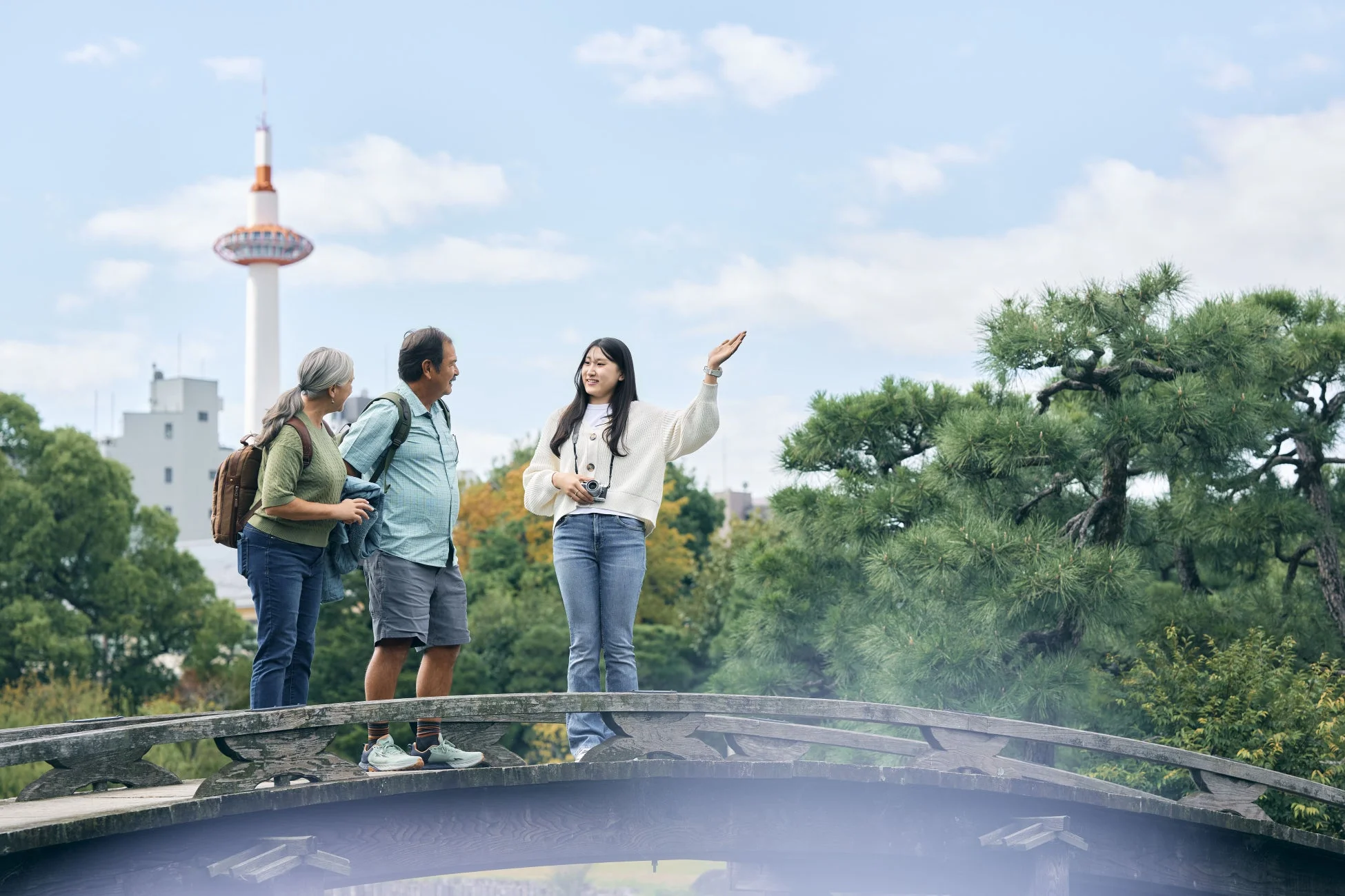 京都タワーを背景に、日本の伝統的な庭園の橋の上で、若い女性がシニアの夫婦に何かを説明している様子です。