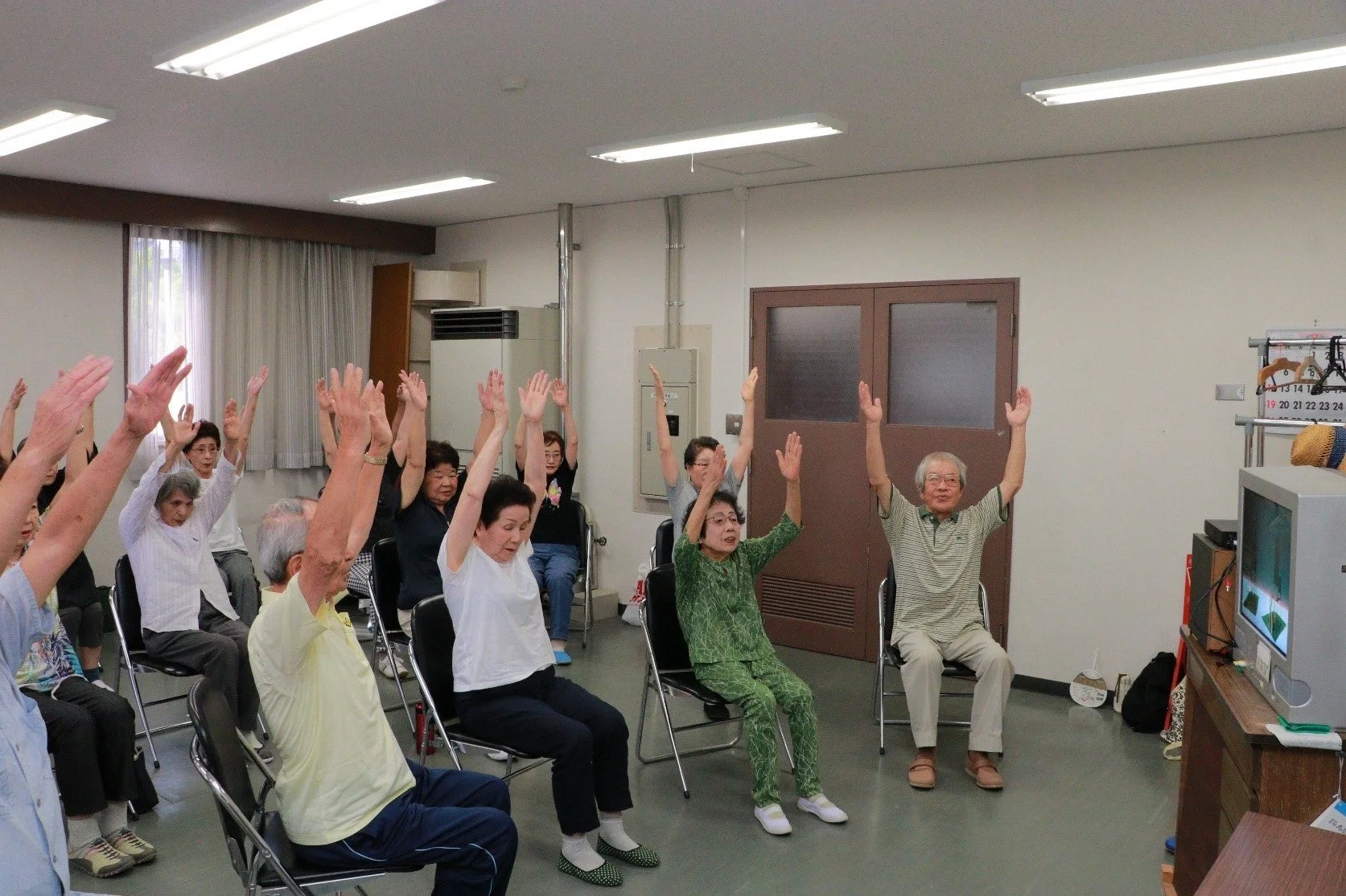 高齢者たちが椅子に座って体操をしている様子。室内で健康維持のためのグループエクササイズが行われていると思われます。