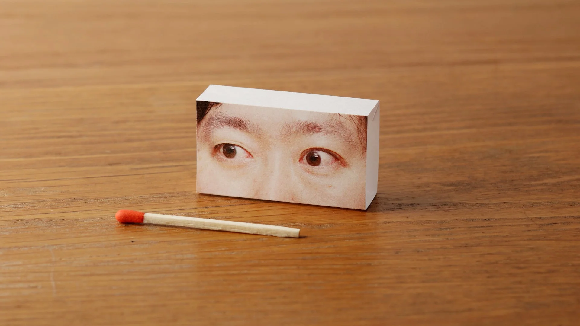 人の目がプリントされた白い箱と一本のマッチ棒が置かれているユニークな静物写真