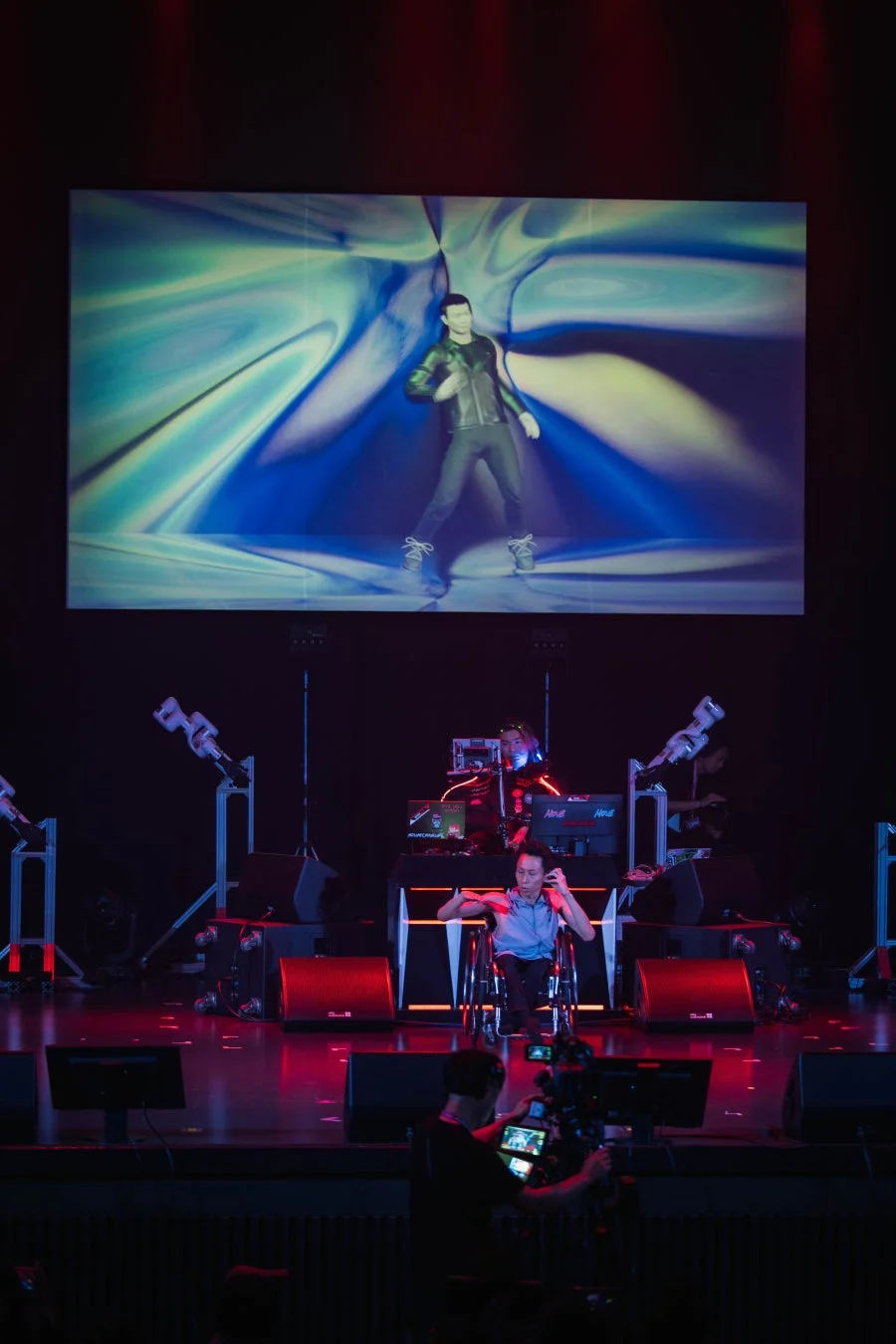 暗いステージで、大型スクリーンに踊る3Dアニメーションが映し出され、車椅子の人物とDJがパフォーマンスを行っている。カメラマンがその様子を撮影しており、テクノロジーとエンターテイメントが融合したライブイベントの瞬間を捉えている。
