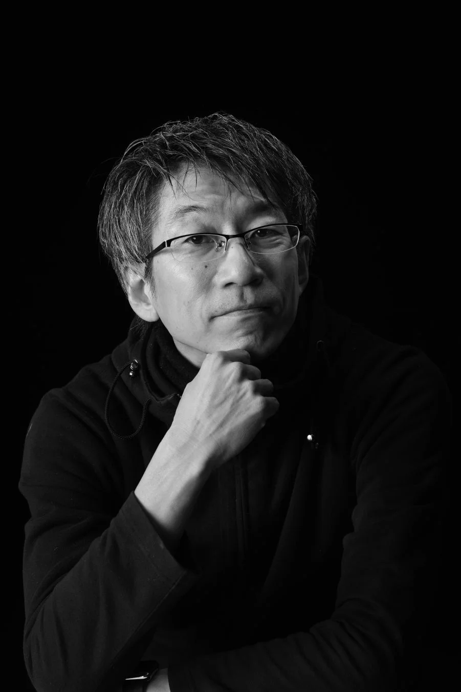 黒い背景の前で、眼鏡をかけた中年男性が顎に手を当ててポーズをとっているモノクロのポートレート