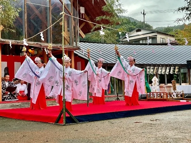 神社らしき場所で、白い着物と緋袴を身につけた若い女性たちが、鈴を持って伝統的な舞を奉納している