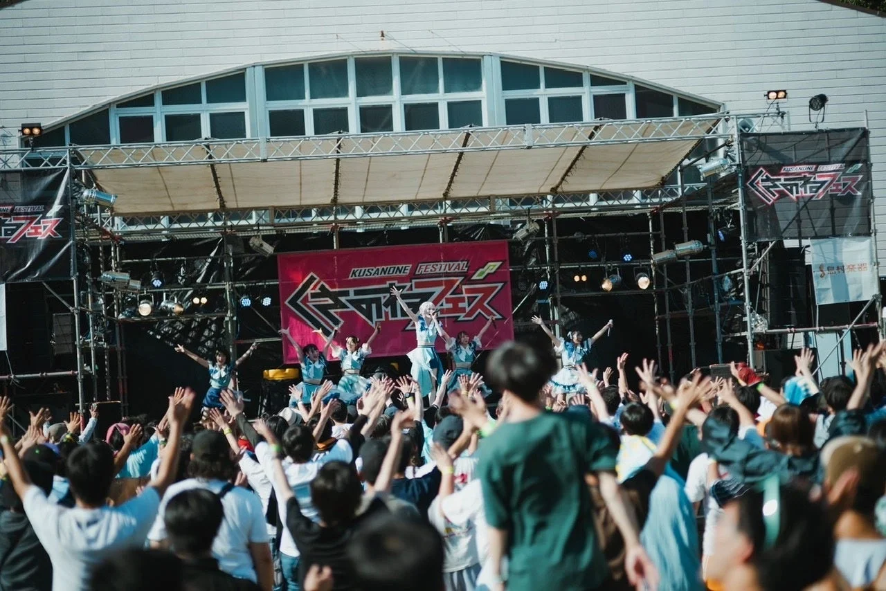 屋外ステージでパフォーマンスを行うアイドルグループと、手を挙げて盛り上がる大勢の観客