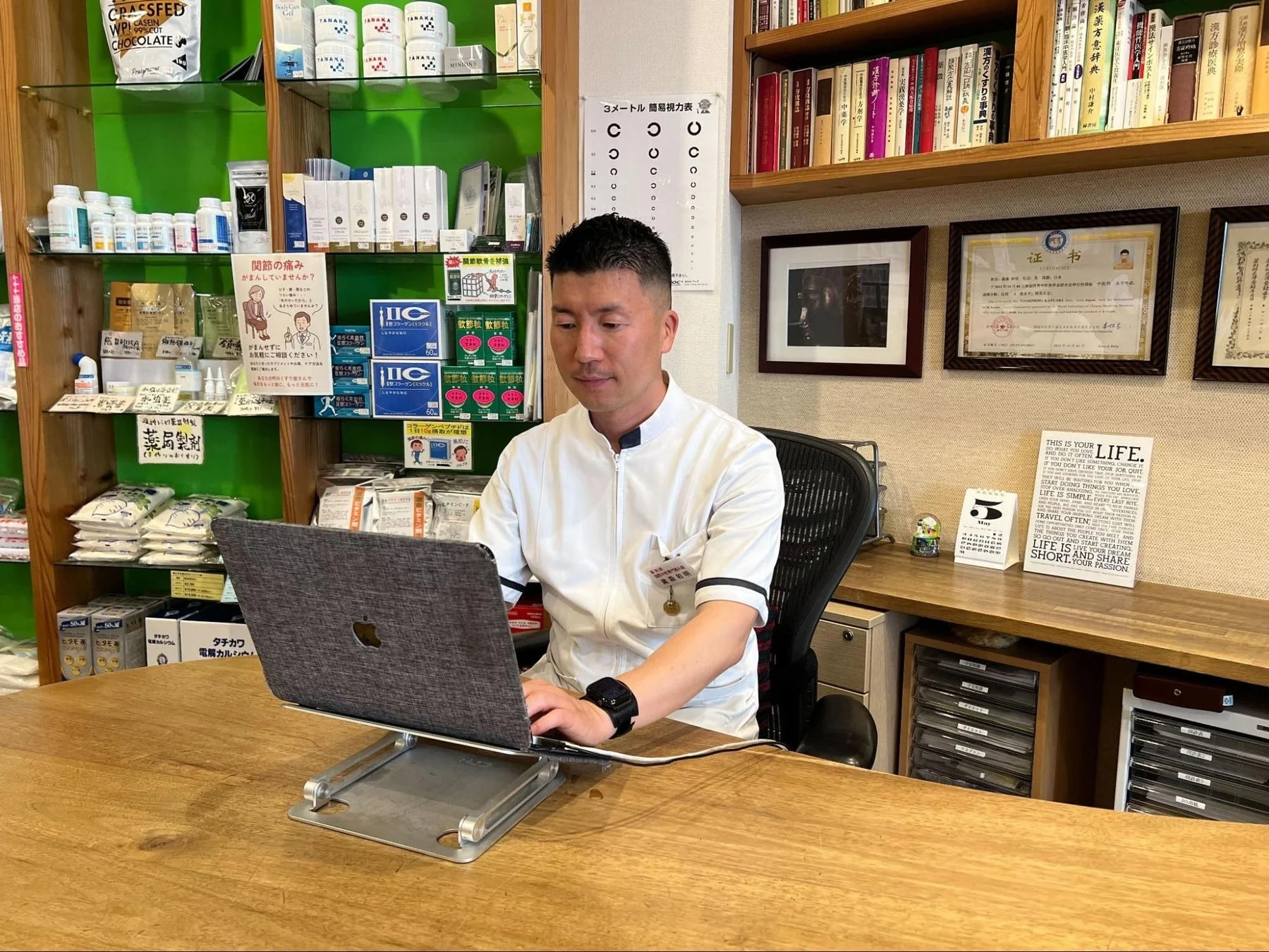 白衣を着た男性が薬局のような場所でノートパソコンを操作しています