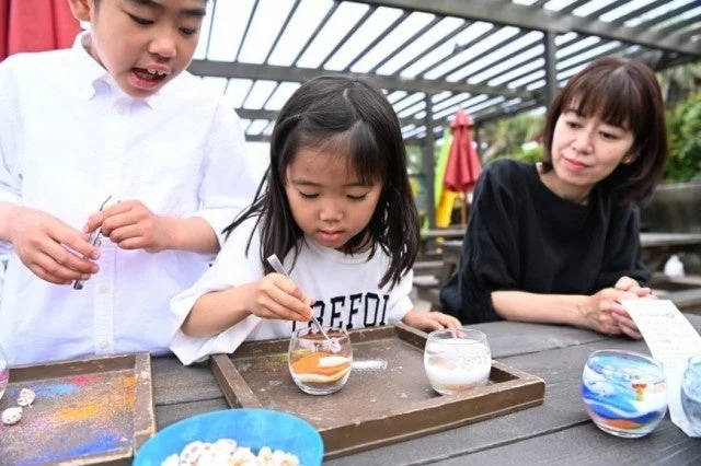屋外で子供たちがサンドアートのようなクラフト体験を楽しんでいます。男の子と女の子が小さなグラスに色のついた素材を熱心に重ねており、隣では女性が優しく見守っています。家族で過ごす楽しい手作りアクティビティの一コマです。