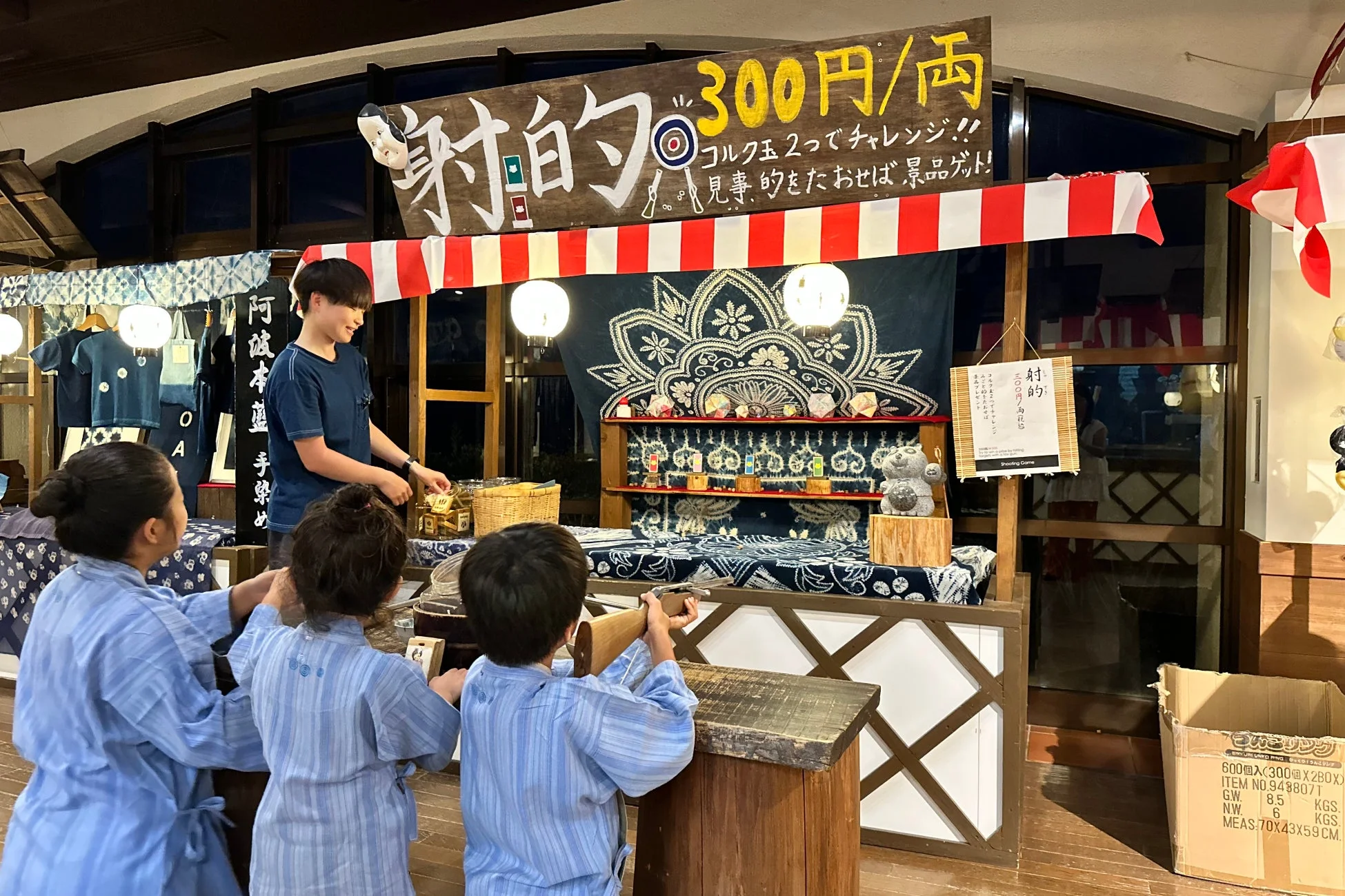 浴衣のような装いの子供たちが射的ゲームを楽しむ様子。縁日のような雰囲気の屋台で、コルク銃を使って景品を狙っている。和風の装飾が施された空間で、日本の伝統的な遊びが体験されている。