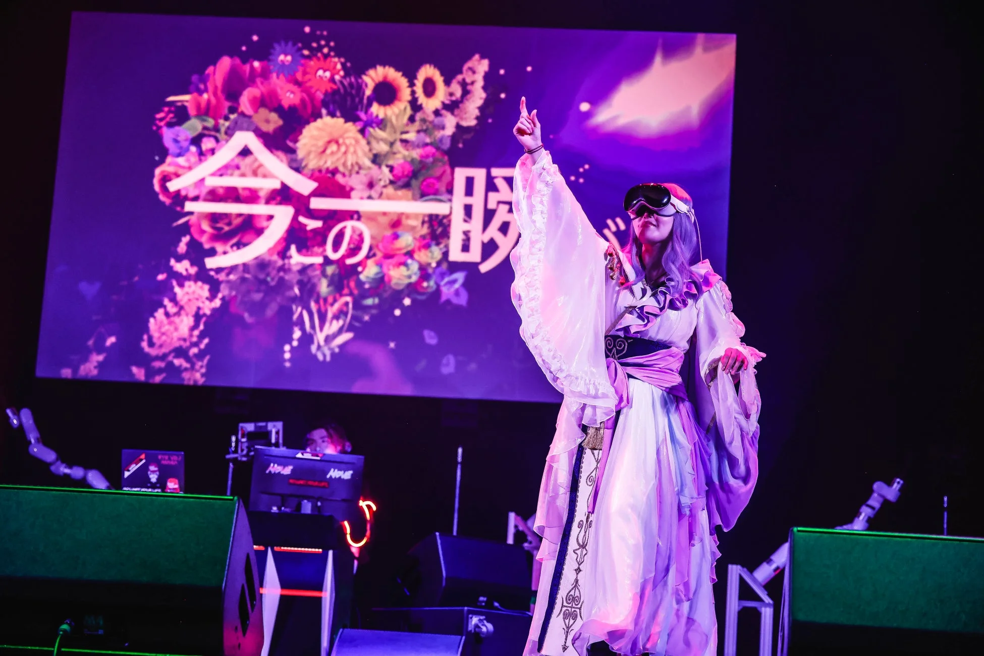 紫色の照明に照らされたステージで、VRゴーグルと豪華な衣装を身につけたパフォーマーが腕を上げて歌唱している。背景の大型スクリーンには、色とりどりの花と「今 この 一瞬」という文字が映し出されており、幻想的なライブパフォーマンスの瞬間を捉えている。
