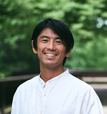 緑豊かな背景を背に、白いシャツを着たアジア人男性がカメラに向かって明るい笑顔を見せているポートレート写真です。親しみやすい表情が特徴的です。