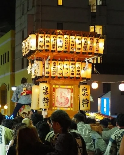 夜の日本の祭りの様子。多数の提灯で飾られた豪華な山車が多くの人々で賑わう通りを進んでいます。