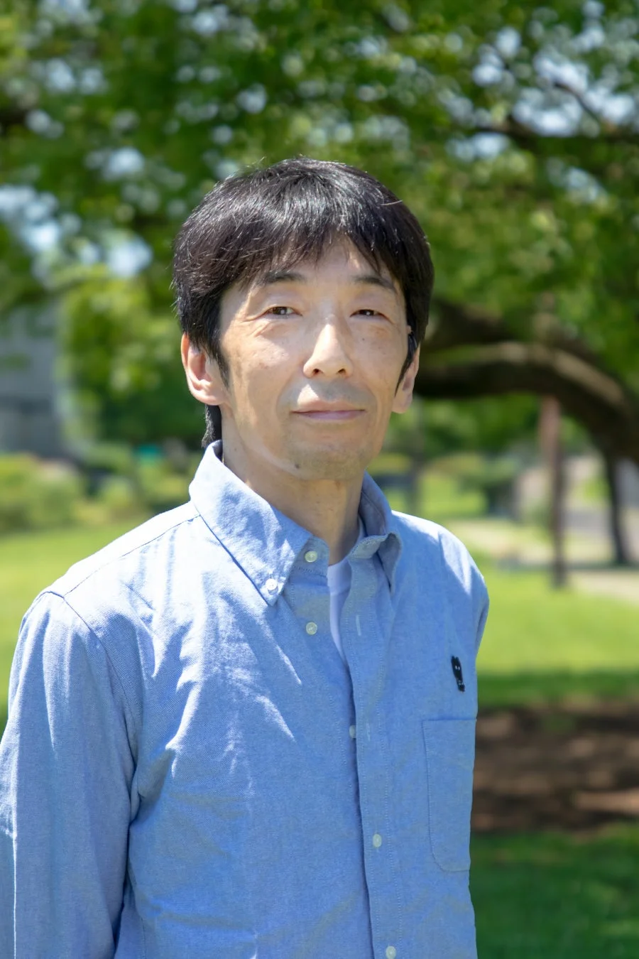 屋外の公園で、青いシャツを着た日本人の男性が穏やかな表情でカメラを見つめているポートレート