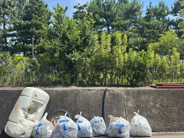 道路脇のコンクリート壁の前に、複数のゴミ袋、大きな発泡スチロールの塊、タイヤなどが放置されている。背景には緑豊かな木々と青空が広がり、不法投棄された廃棄物の状況を示す光景です。