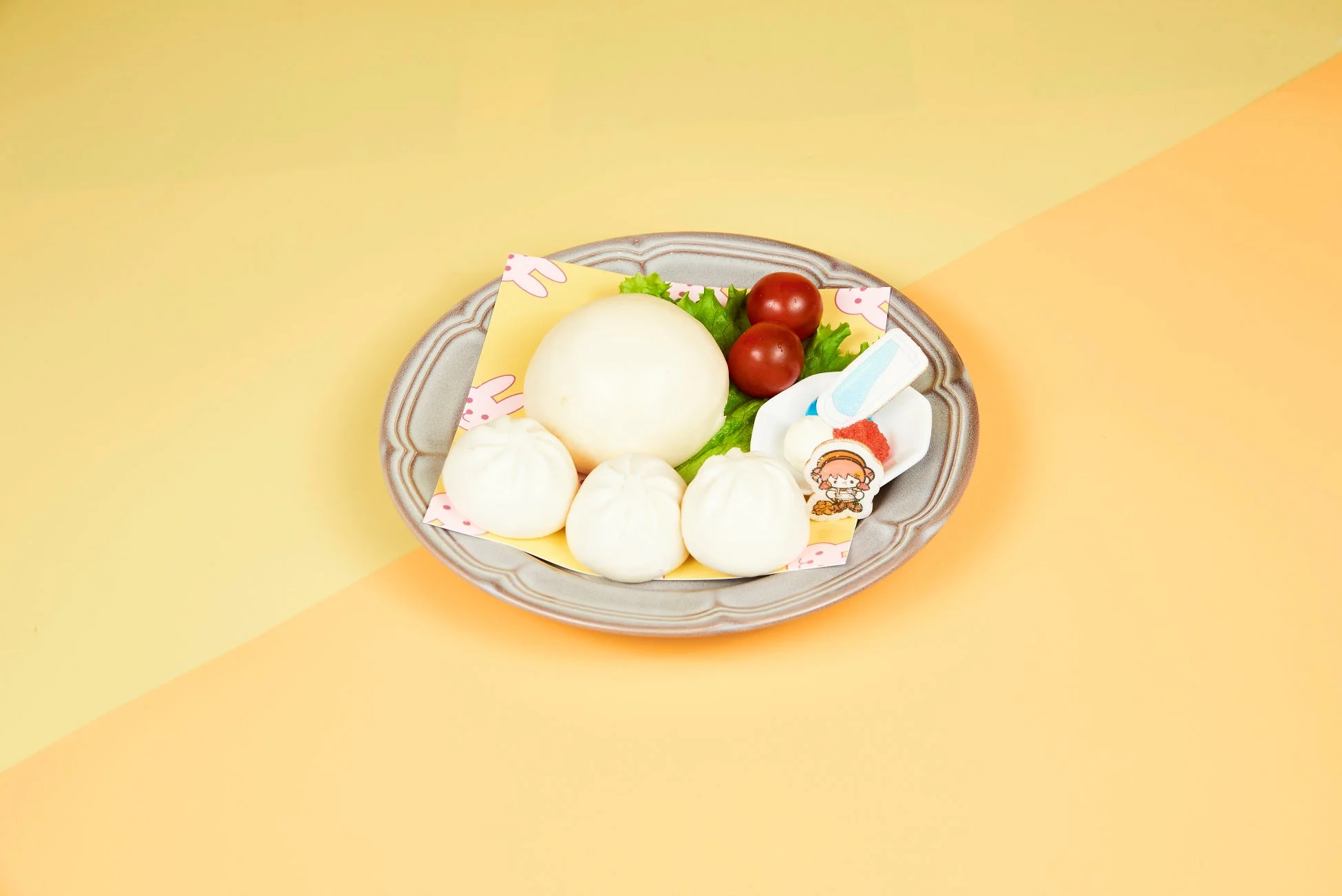 黄色とオレンジ色の背景に置かれたグレーの皿に、大きな白い蒸しパンと小さな蒸しパンが3つ、ミニトマト2個、レタスが盛り付けられています。うさぎ柄のシートが敷かれ、小さな器には赤いトッピングとキャラクターの飾りが添えられています。