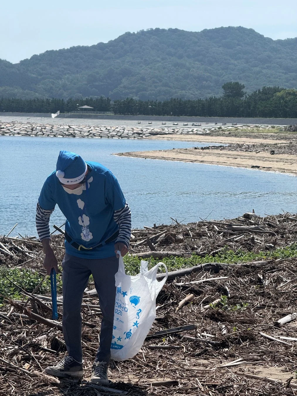 青いサンタの衣装を着た男性が、流木やゴミが散乱する海岸でごみ拾いをしている様子。海と山が広がる自然豊かな場所での清掃活動で、環境保護への取り組みが伺えます。