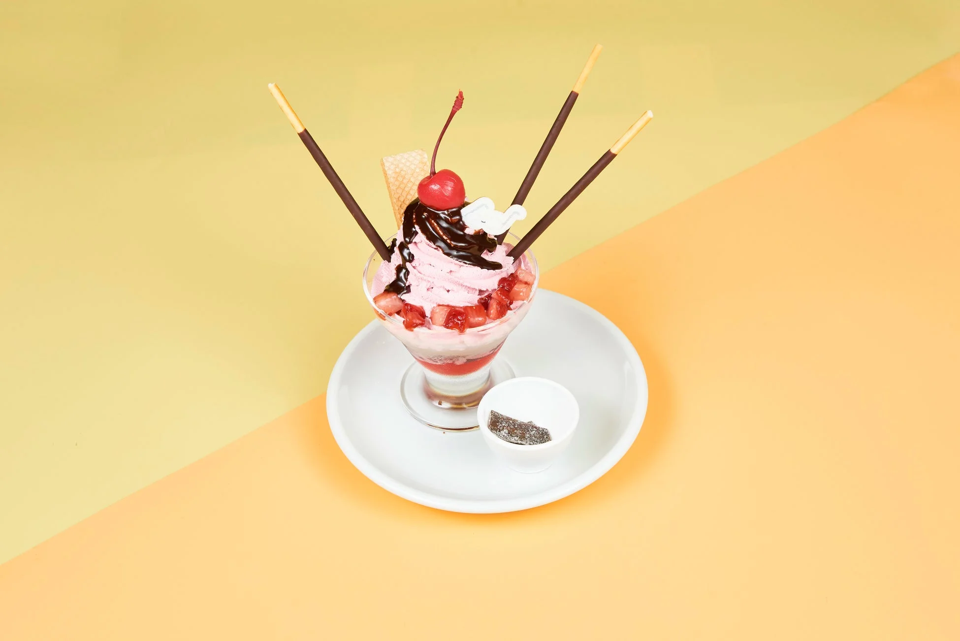 ピンクのいちごアイスクリームとカットしたいちごが層になったパフェです。チョコレートソース、チェリー、チョコレートスティック、ウエハース、象の飾りでデコレーションされており、白い皿に置かれ、背景は黄色とオレンジに分かれています。