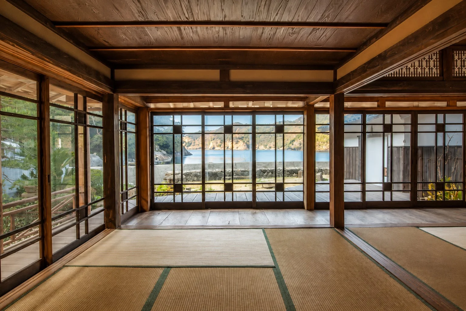 畳が敷かれた伝統的な和室から、大きな窓越しに穏やかな海と山々が広がる絶景を望む。木造建築が趣深く、静かで落ち着いた雰囲気を醸し出している。