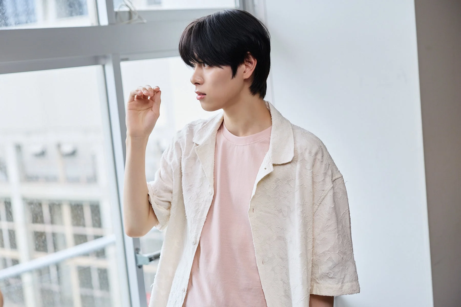 黒髪の若い男性が、ピンクのTシャツと淡い色の半袖シャツを着用し、窓辺で物思いにふけるように横を向いているポートレート写真。