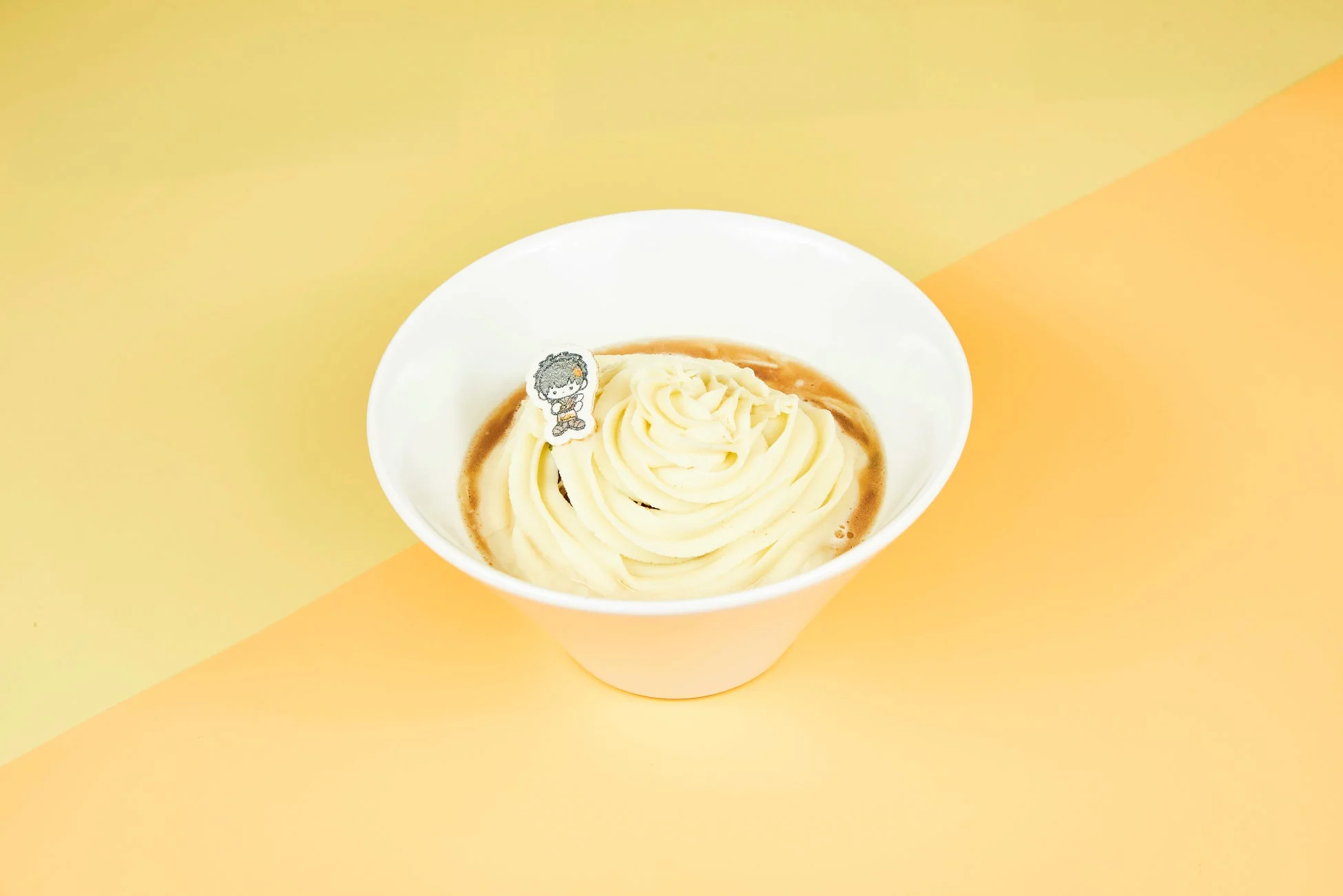 白いボウルに入った、クリーム状の料理が写っています。茶色いソースの上に、絞り出されたような白いクリームが盛り付けられ、小さなアニメキャラクターのピックが添えられています。背景は黄色とオレンジ色のツートンカラーです。