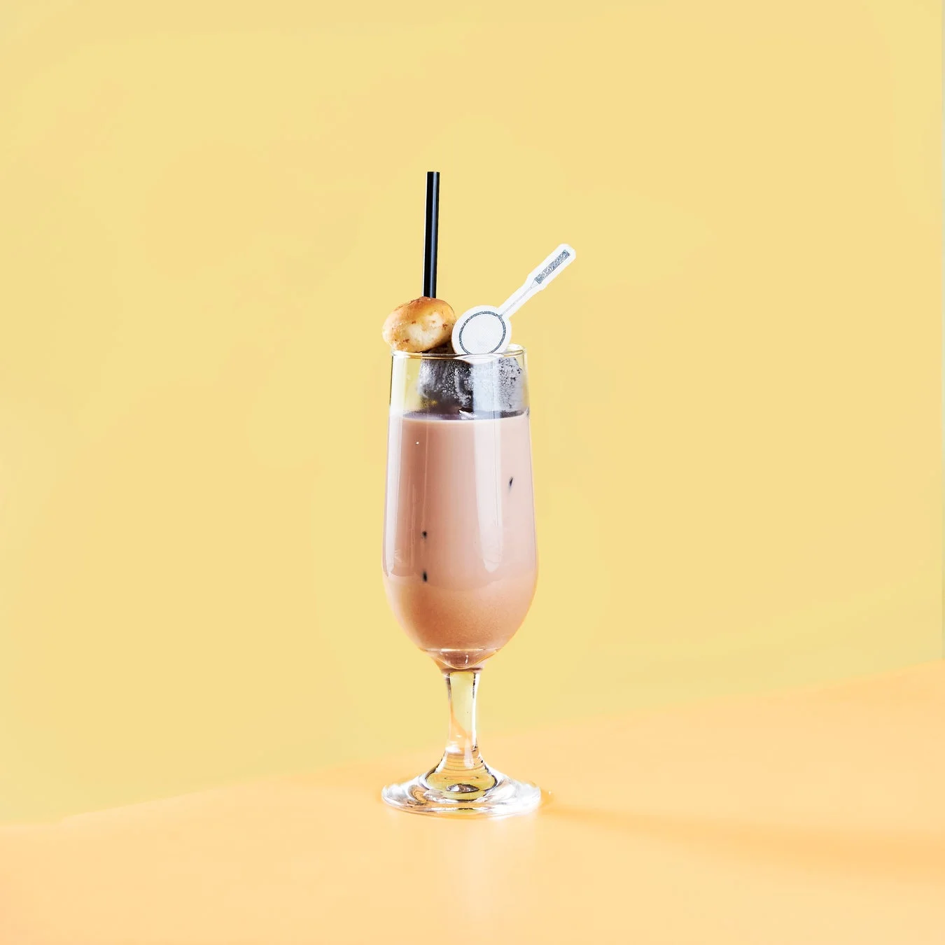 黄色い背景に置かれた、アイスが入った茶色い飲み物のグラス。黒いストローと小さな丸いお菓子、装飾的なスプーンが添えられており、シンプルながらも魅力的なデザートドリンクの画像です。