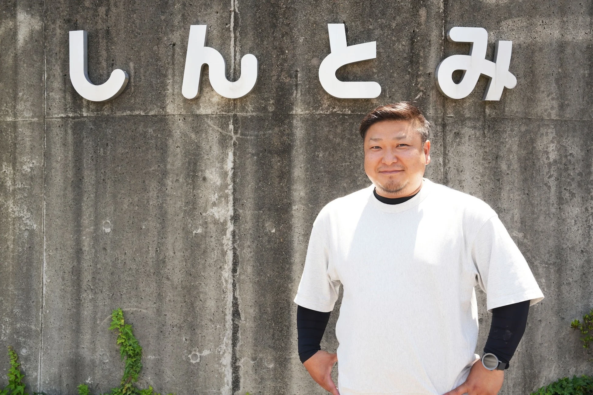 「しんとみ」と書かれたコンクリートの壁の前に立つ、笑顔の男性のポートレート写真です。