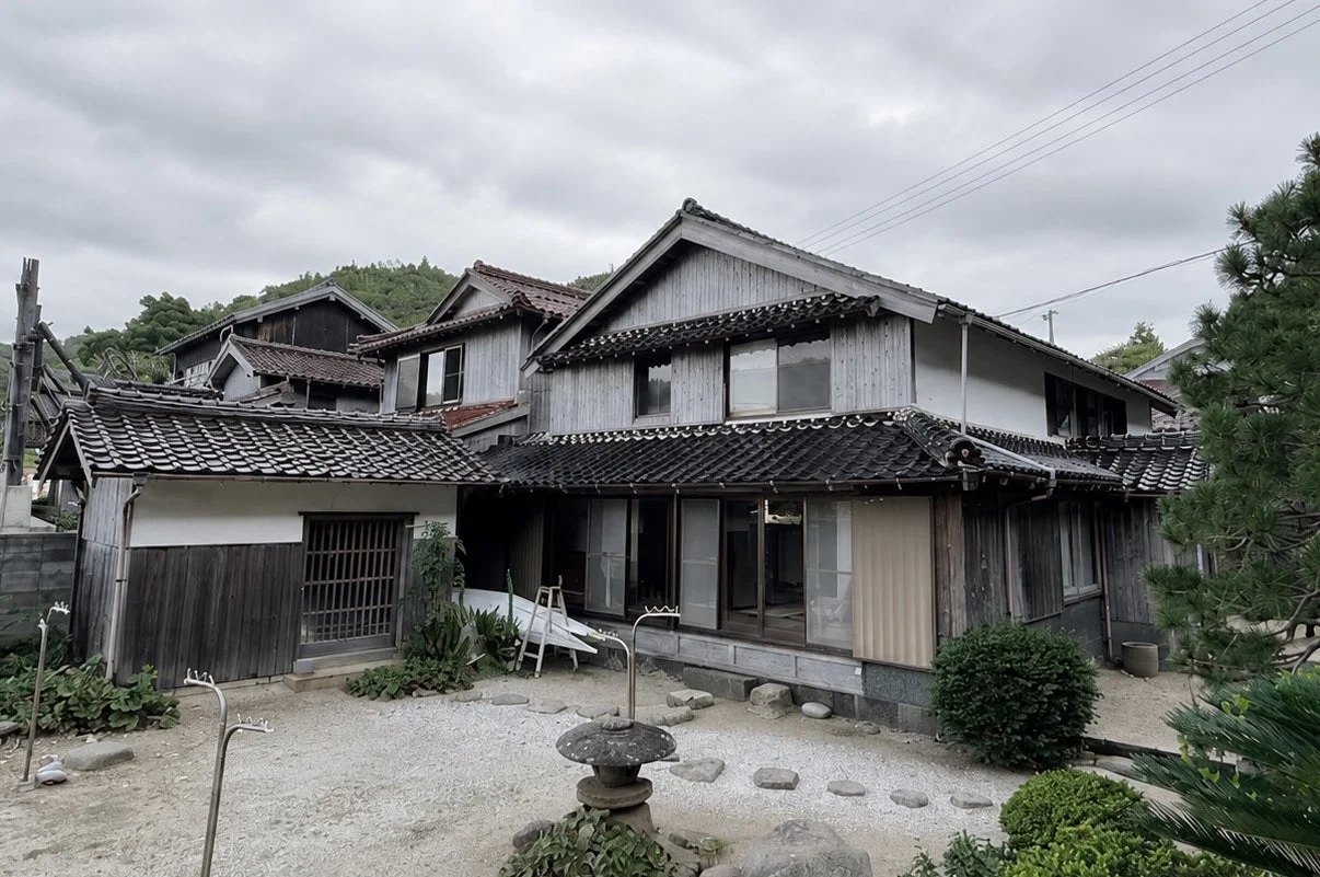 瓦屋根と木造の伝統的な日本家屋が立ち並び、手前には石灯籠のある砂利の庭が広がる風景。曇り空の下、落ち着いた日本の田舎の雰囲気を醸し出しています。